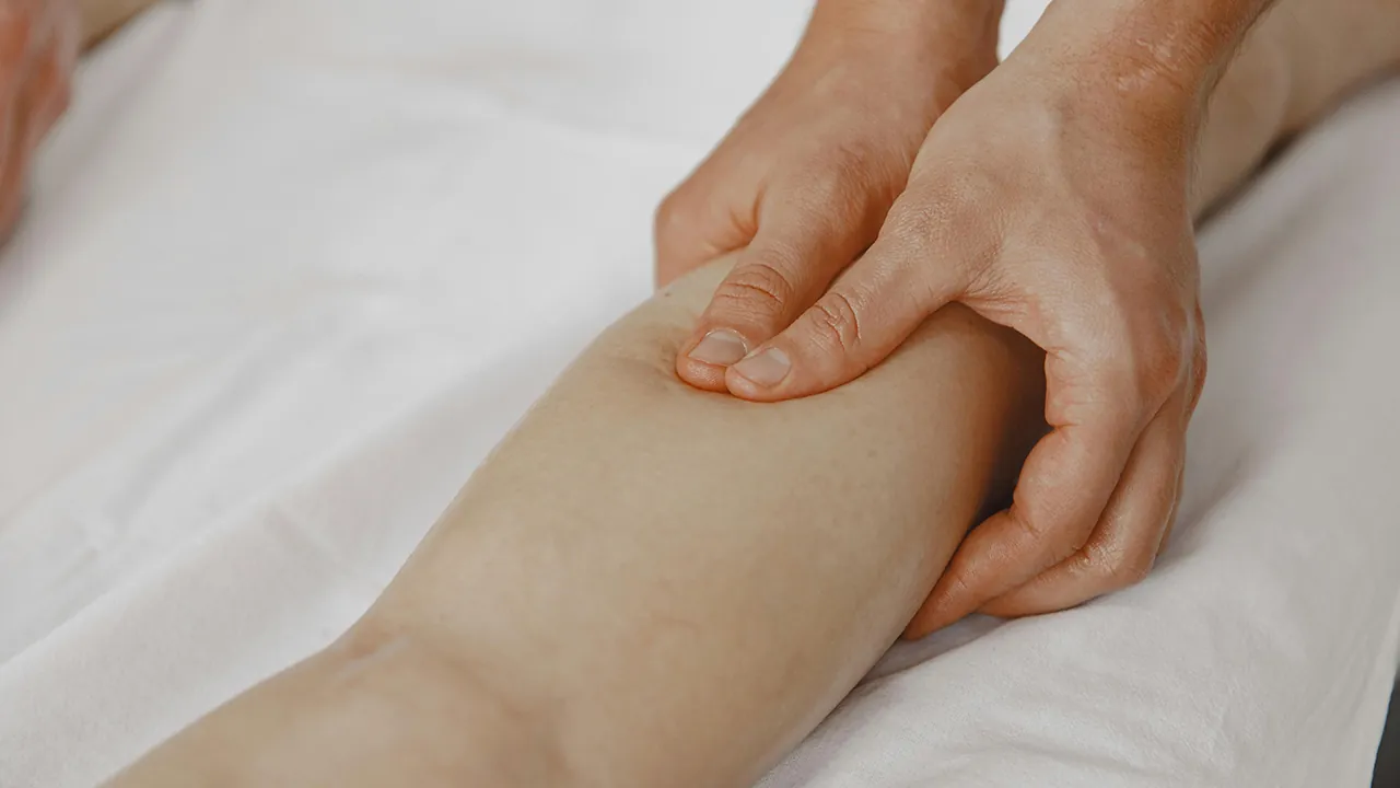 A masszőr két kézzel masszírozza a fehér lepedőn pihenő lábszárat